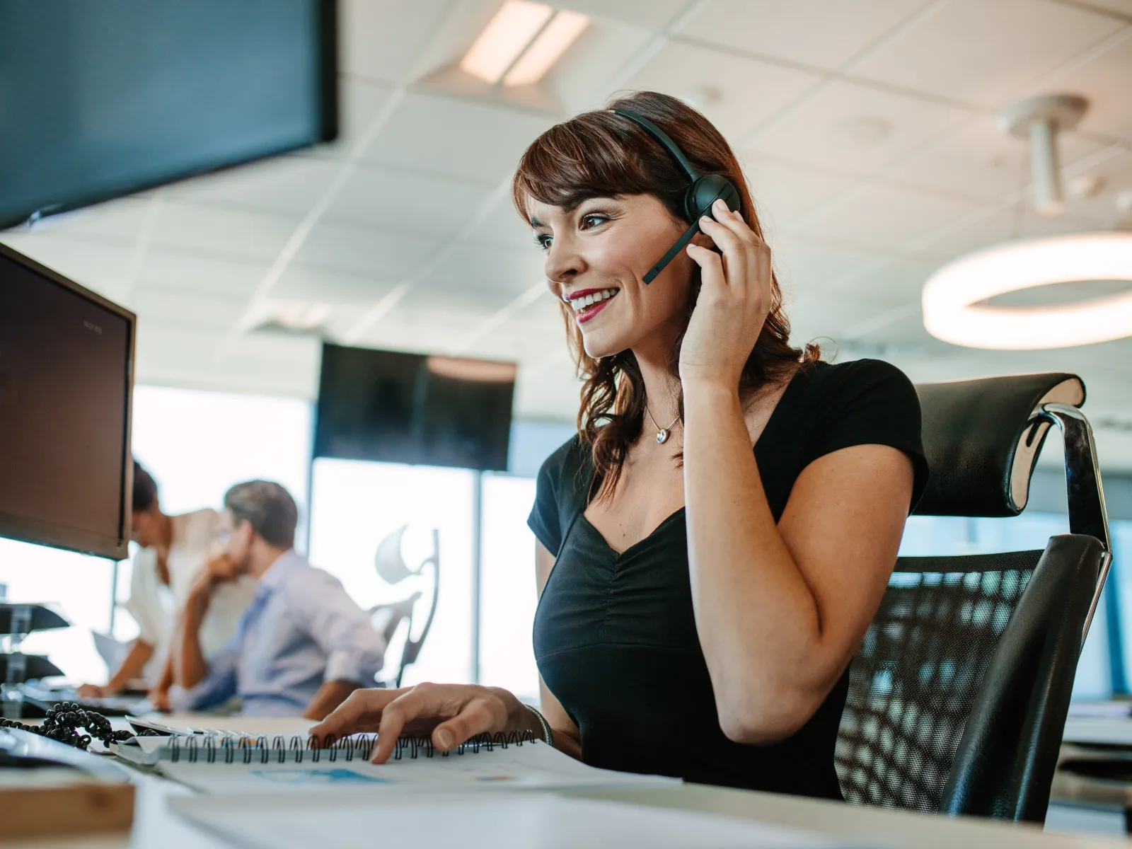 Smiling woman wearing headset working at computer in modern office call center environment.