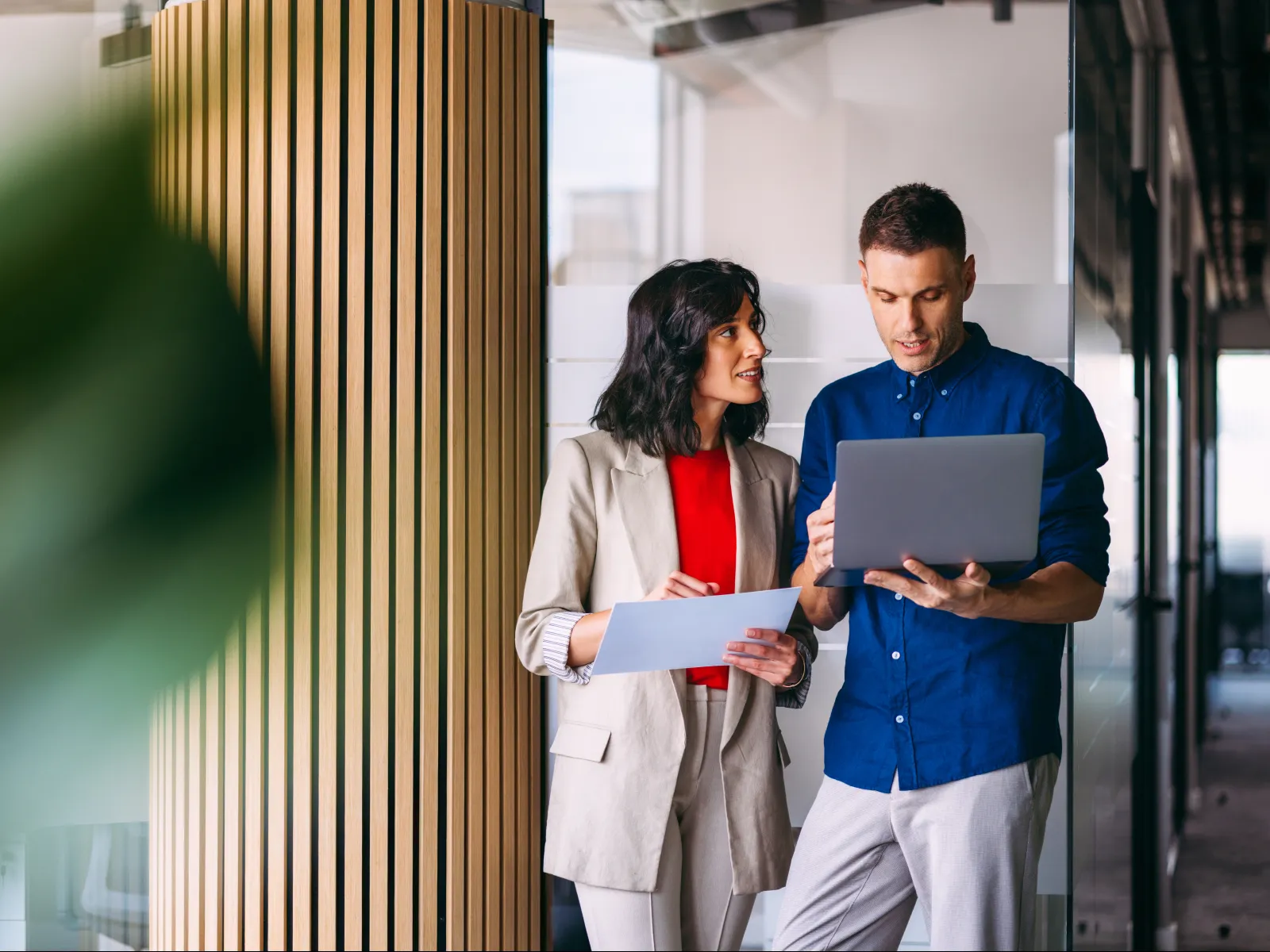 Two coworkers discussing work with a laptop and documents in a modern office space with wooden paneling.