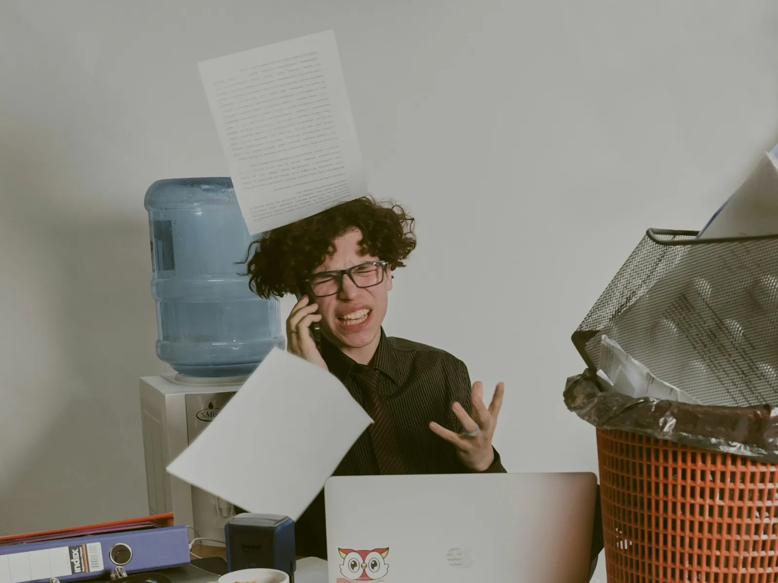 Frustrated person at cluttered desk with papers flying around and a laptop, expressing stress or overwhelm.