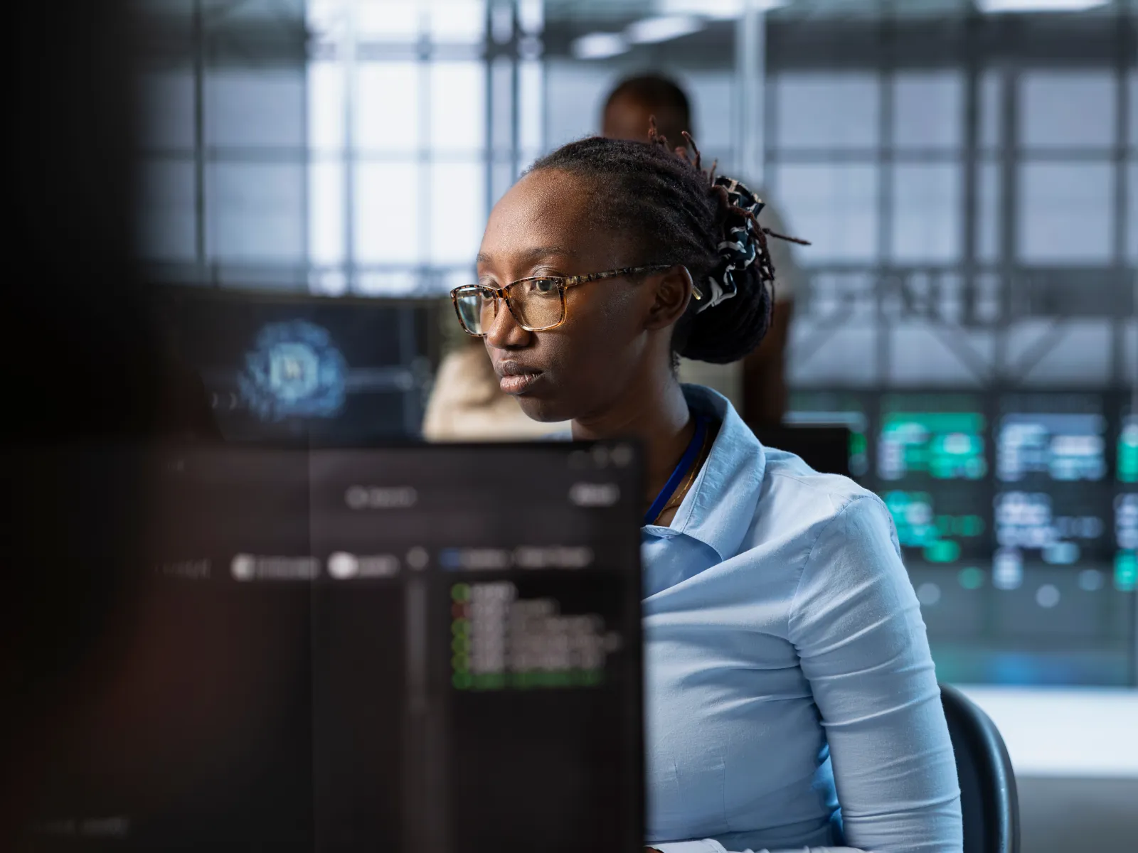 Focused woman wearing glasses working on computer in modern office with digital screens in the background