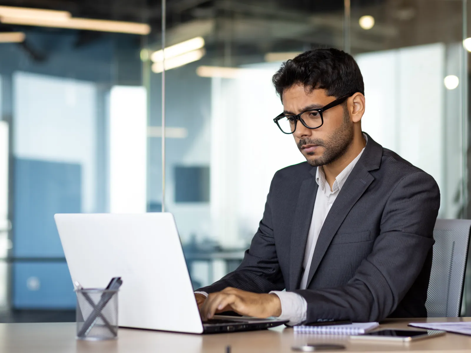 Professional man in glasses working on laptop in modern office with glass walls and natural light.