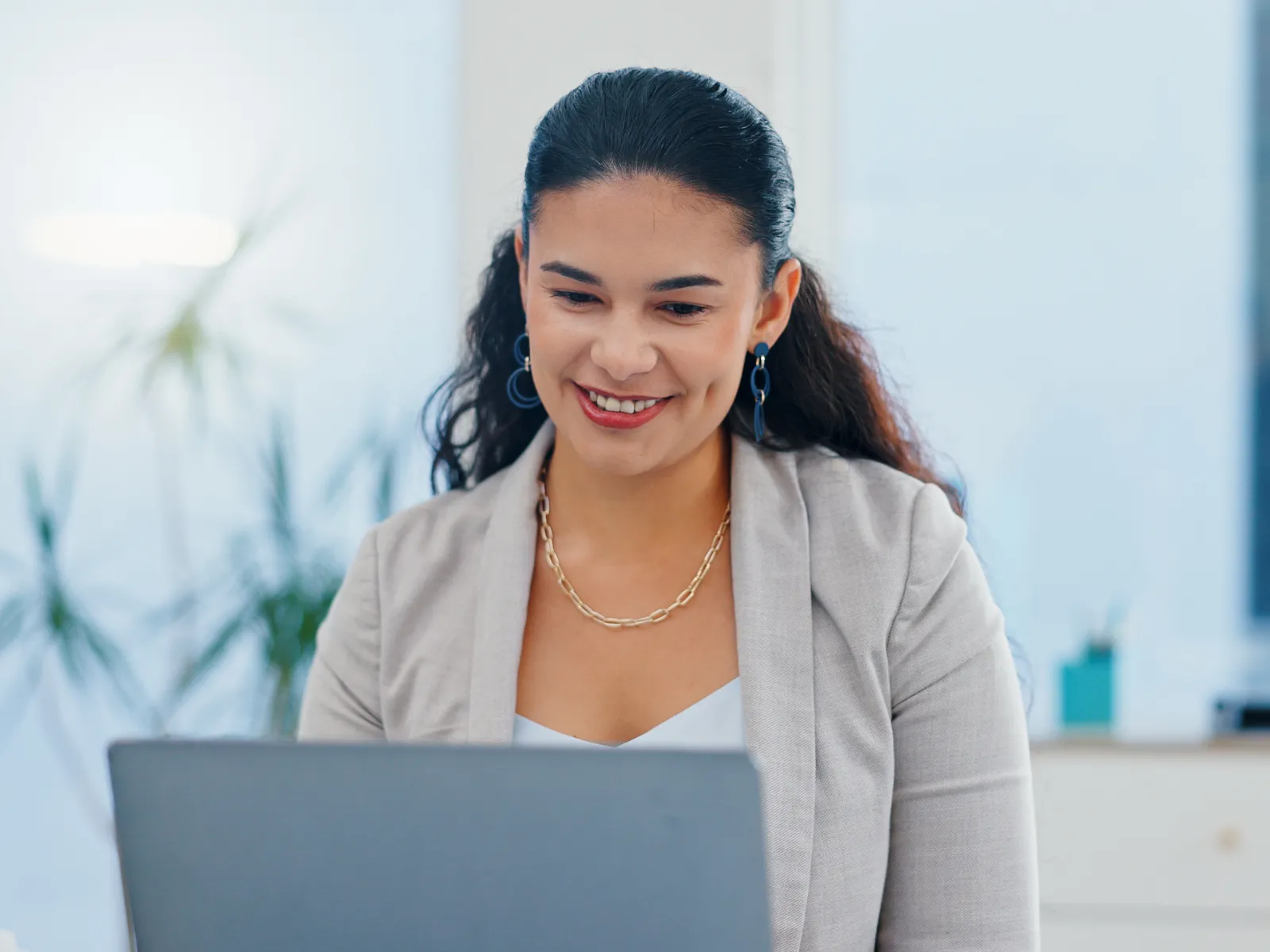 Smiling businesswoman in blazer working on laptop in bright modern office with plants in background