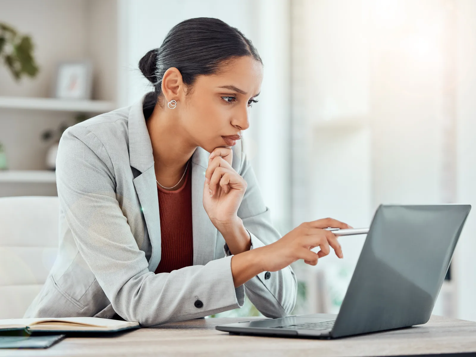 Businesswoman in gray blazer focused on laptop screen pointing at it while sitting at office desk