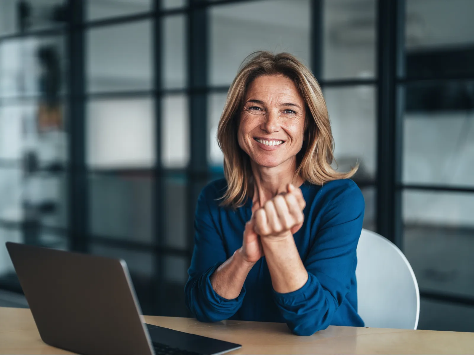 Smiling woman in blue blouse sitting at desk with laptop in modern office with glass partitions.
