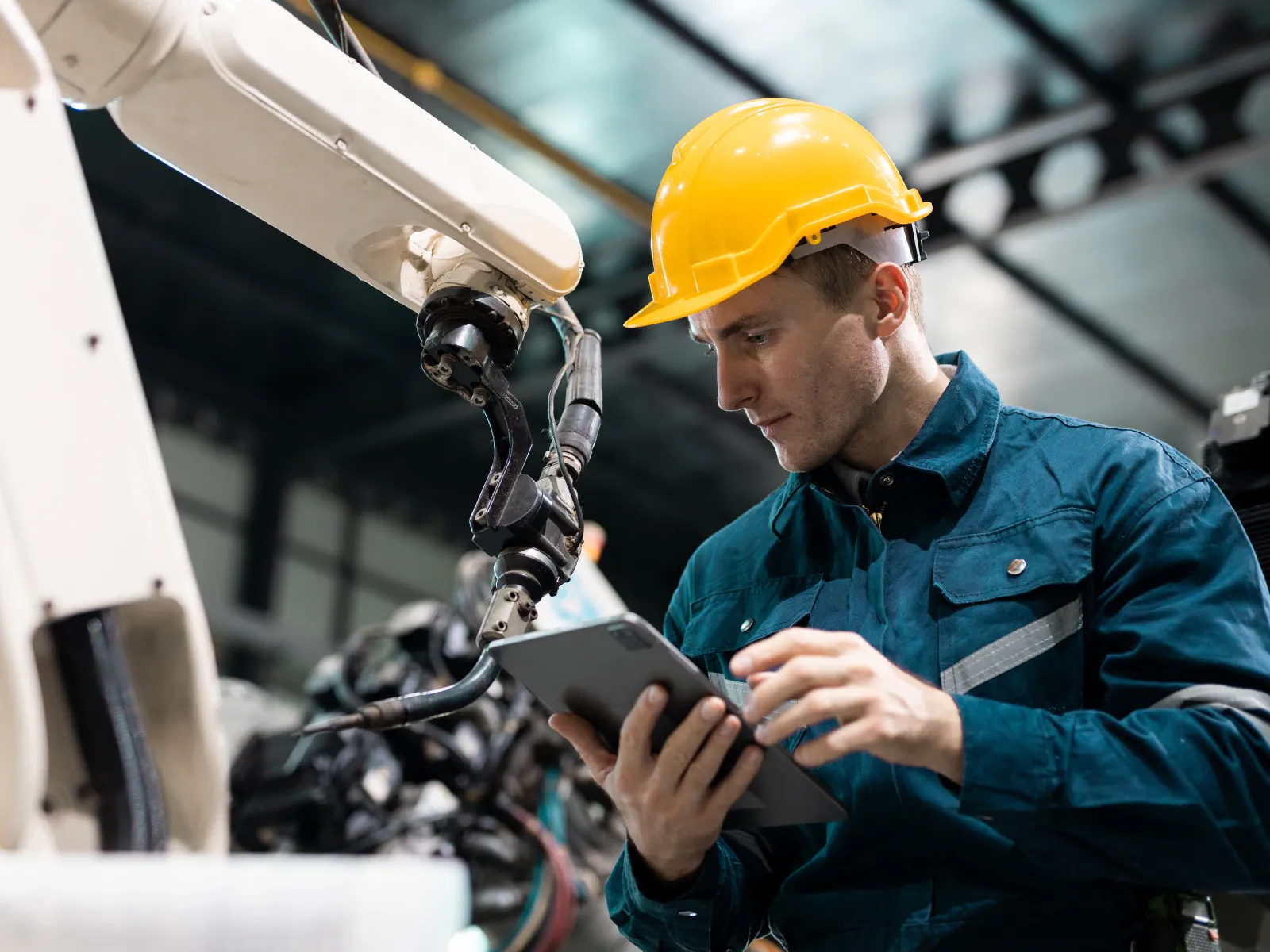 Engineer in a yellow helmet using a tablet to operate robotic arm in an industrial manufacturing setting