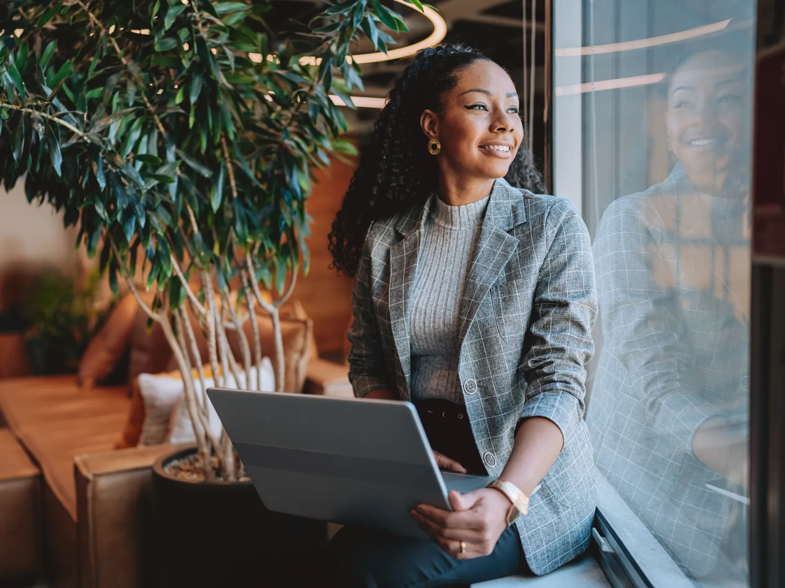 Smiling woman in gray blazer holding laptop and looking out window in modern office with plants and wood decor