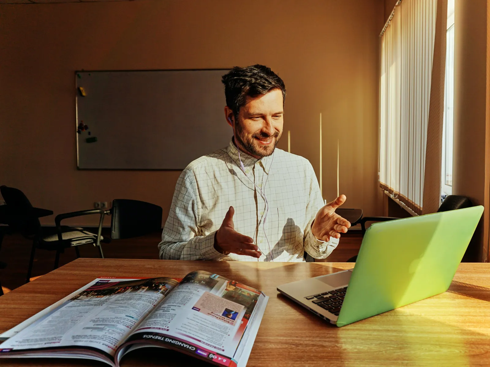 Man smiling and gesturing while video calling on laptop in a sunlit office with an open magazine on the desk.
