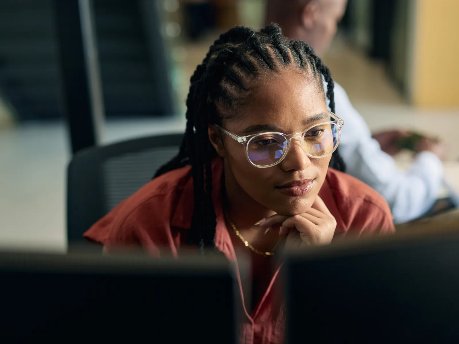 Young woman with glasses and braided hair focused on dual monitors in a modern office setting.