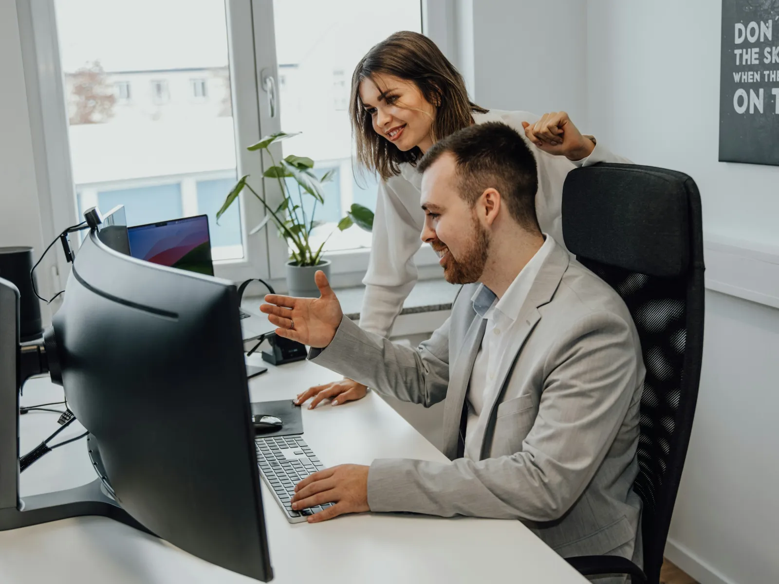 Two colleagues collaborating at a desk with dual monitors in a bright modern office space.