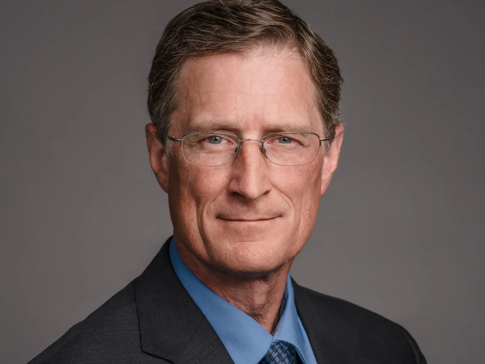 Professional portrait of a mature man in a suit with glasses, featuring a neutral background and a confident expression.