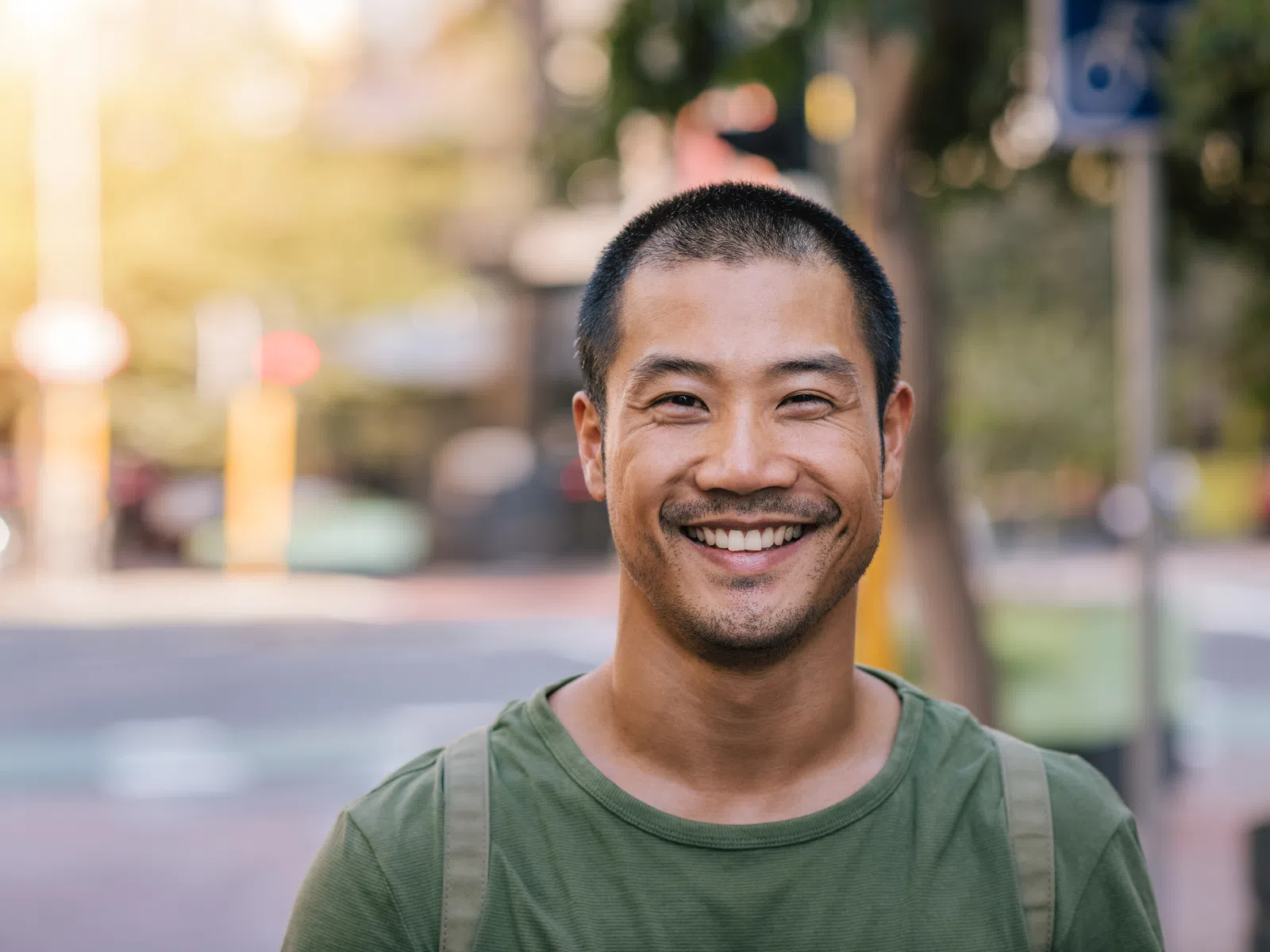 Smiling Asian man with short hair wearing a green shirt and backpack outdoors in a city setting.