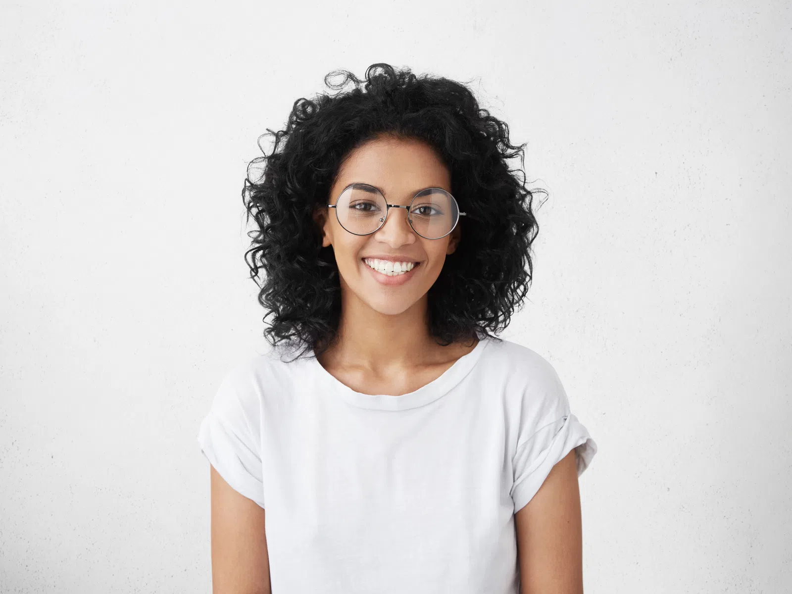 Smiling young woman with curly hair and glasses wearing a white t-shirt against a plain white background