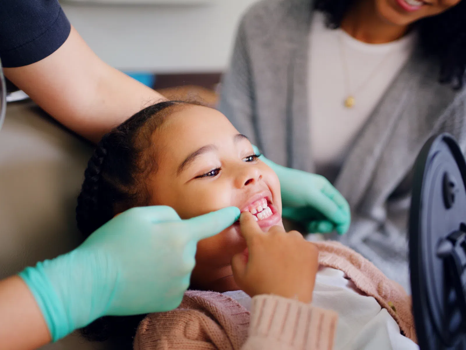 Child smiling while dentist with gloves examines teeth, mother holding mirror in dental clinic.