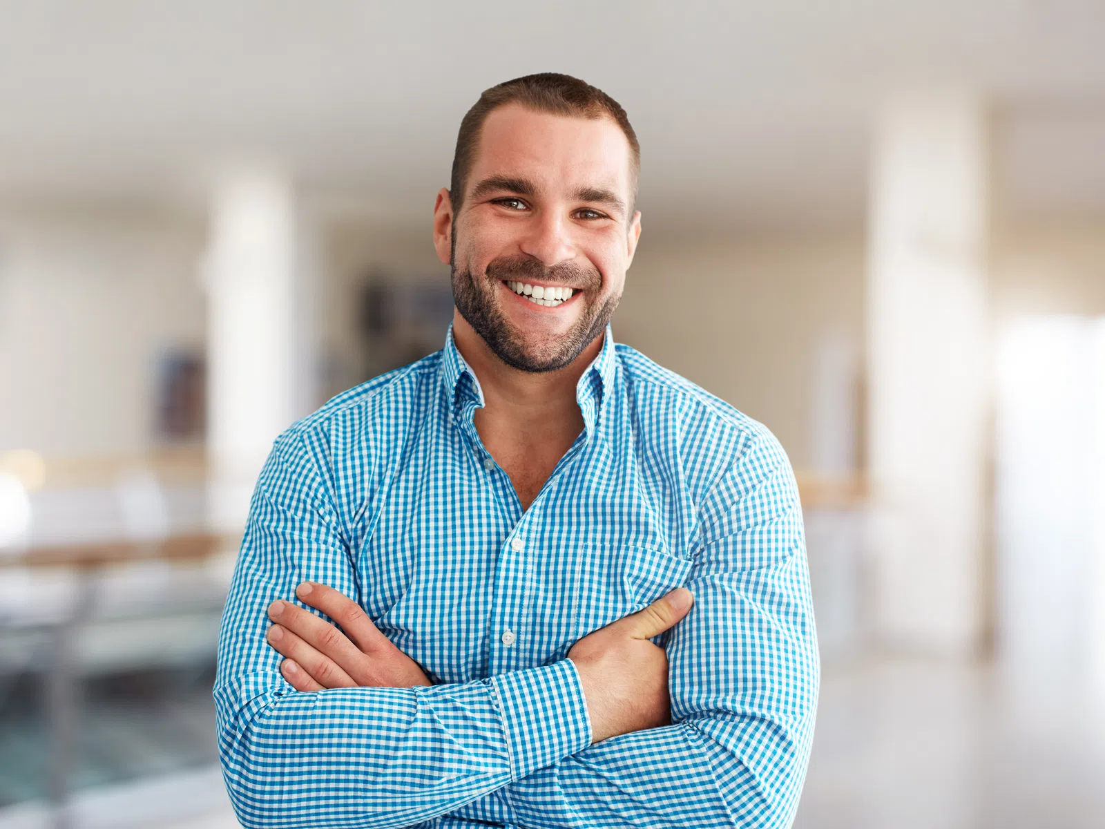 Smiling man in blue checkered shirt with crossed arms standing in bright modern office space.