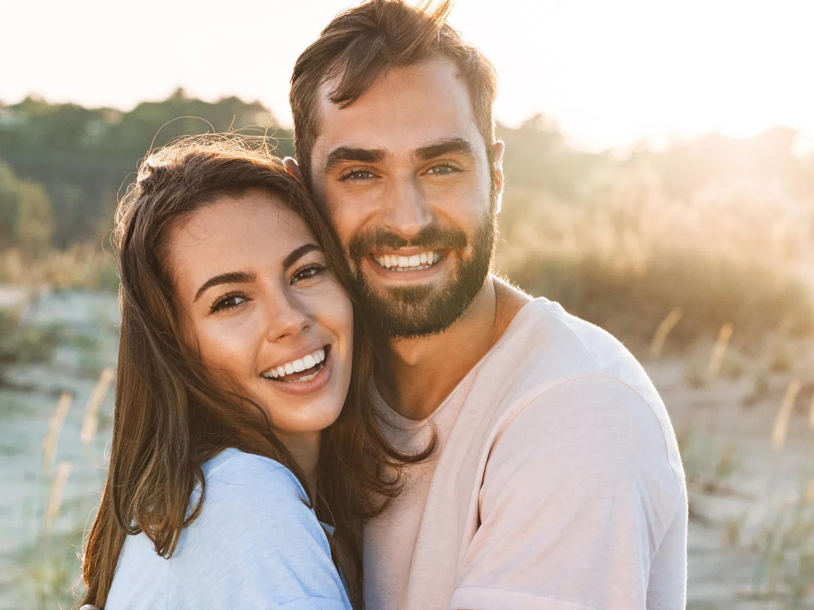 Smiling young couple embracing outdoors in natural sunlight with greenery and sandy ground in the background