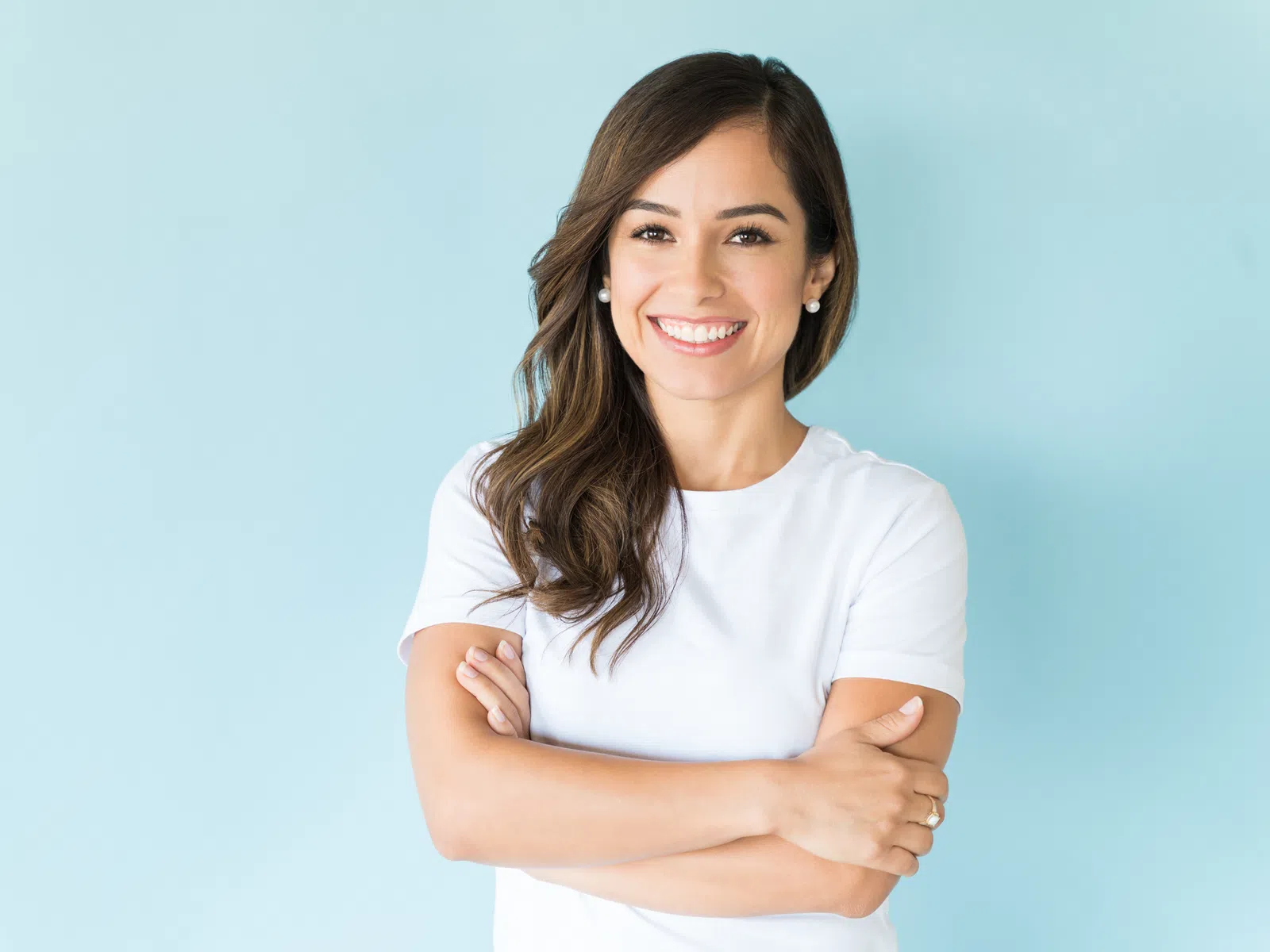 Smiling woman with long brown hair in white shirt stands with arms crossed against light blue background