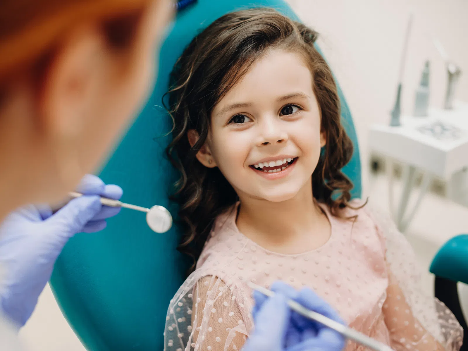 Smiling young girl sitting in dental chair during checkup with dentist holding dental mirror and explorer.