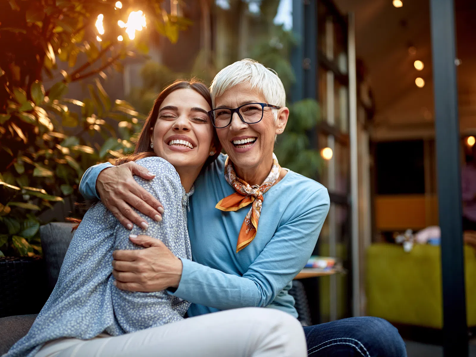 Older woman and young woman smiling and hugging outdoors in a cozy café setting with warm lighting.