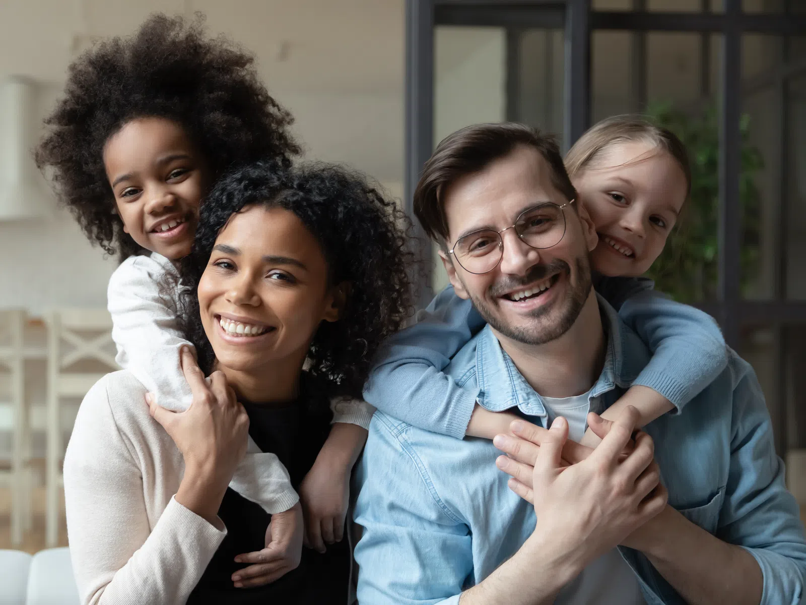 Happy mixed-race family with parents and children smiling and hugging indoors in a bright living room.