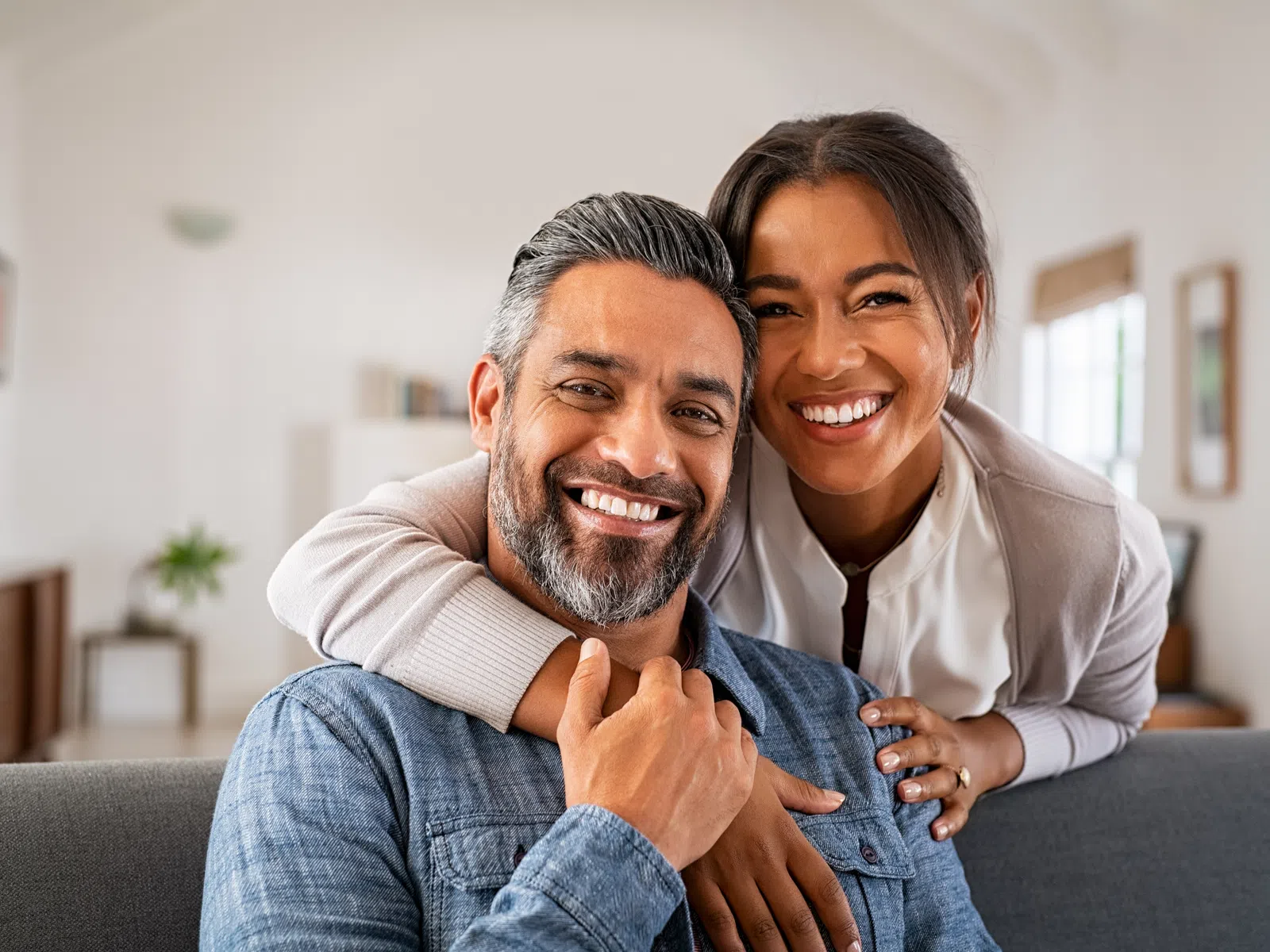 Happy couple smiling and hugging on a couch in a bright living room with cozy decor and natural light.
