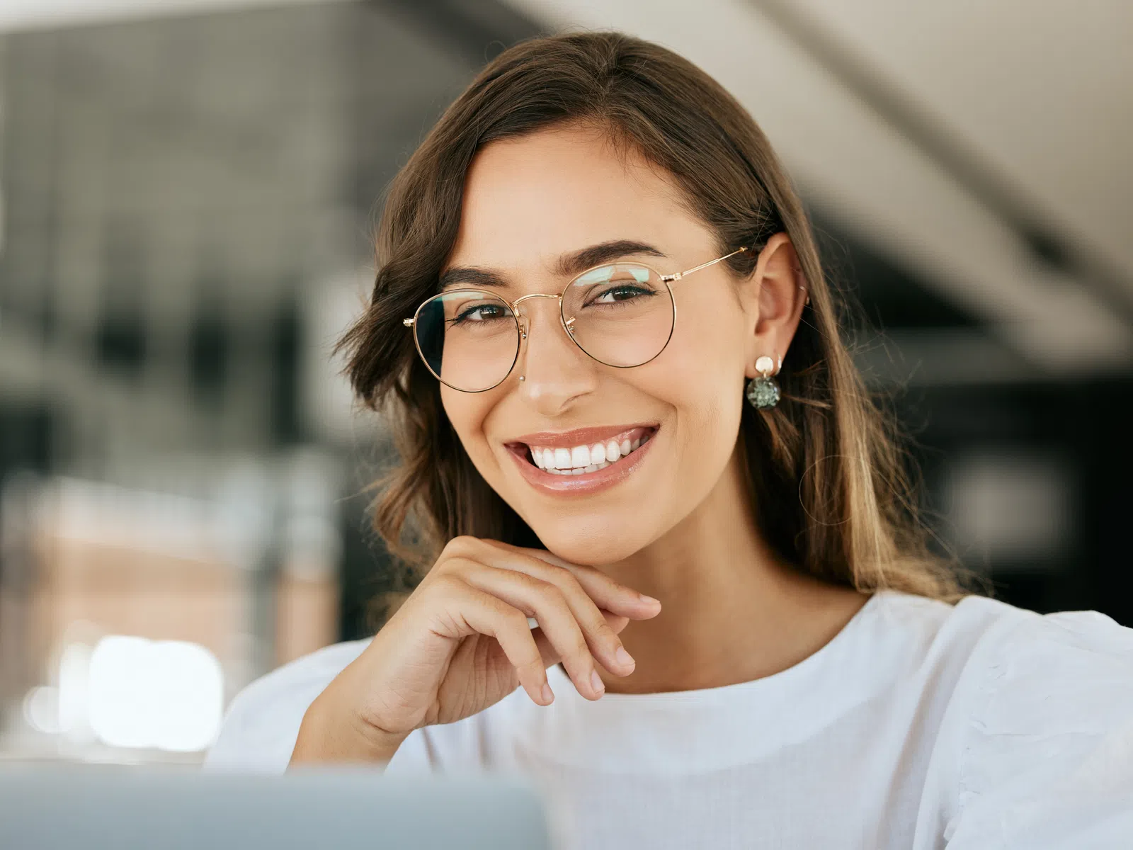 Smiling woman with glasses and pearl earrings sitting indoors with hand near chin, wearing white blouse