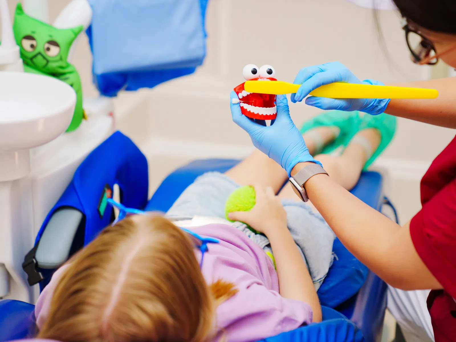 Dentist demonstrates proper brushing on a toy mouth to a child patient lying in the dental chair.