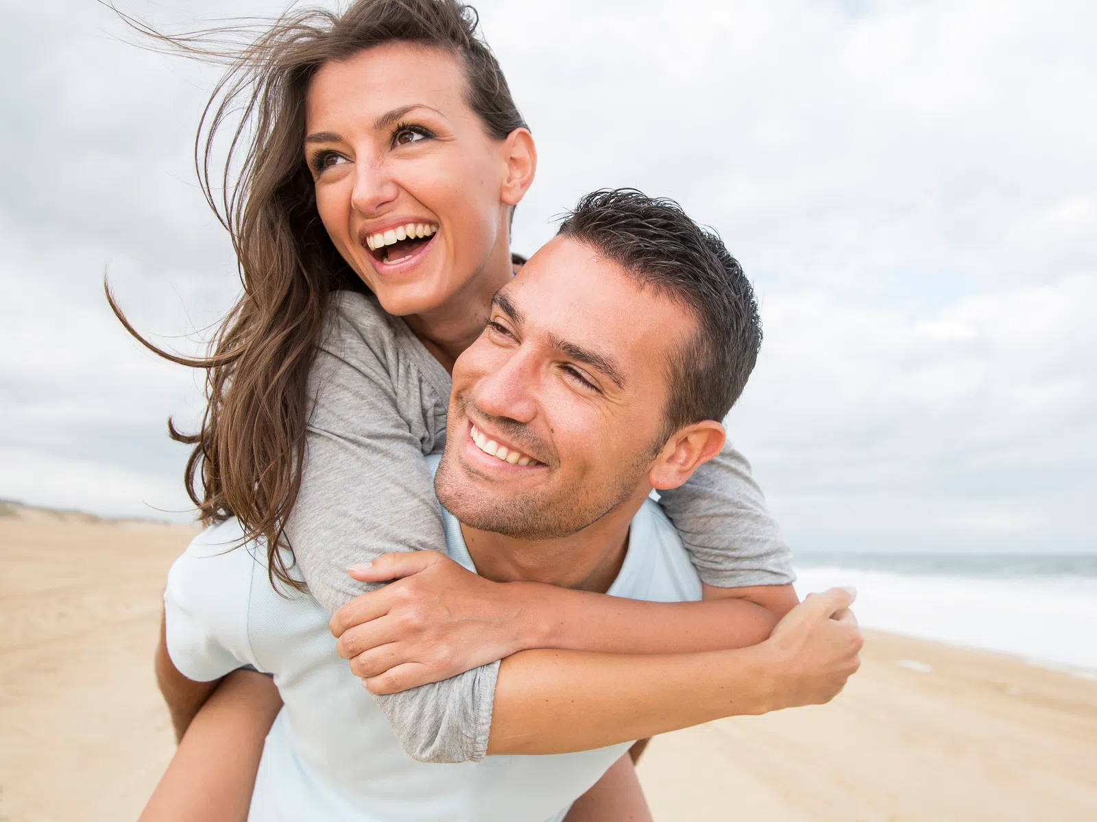 Happy couple enjoying a piggyback ride on a sandy beach with ocean and cloudy sky in the background