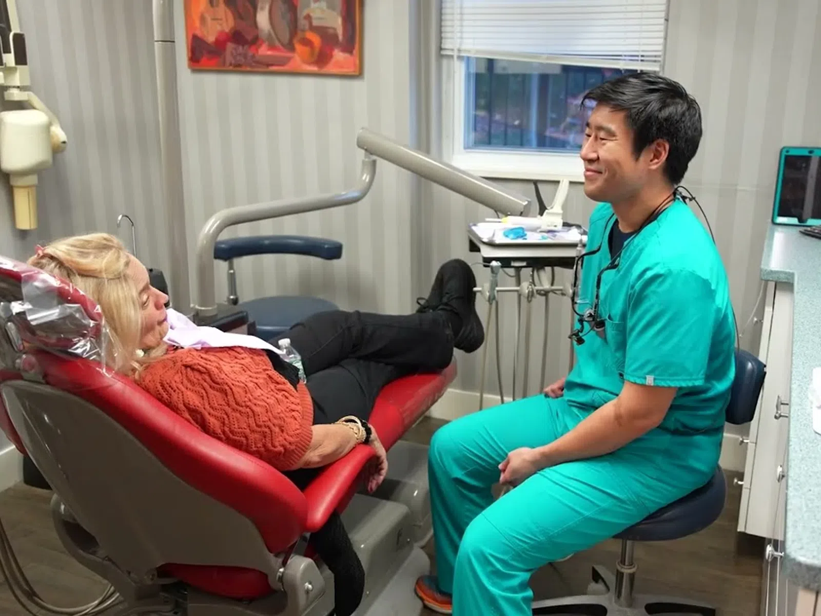 Dentist in green scrubs talking with a patient reclining in a red dental chair in a clinic room.