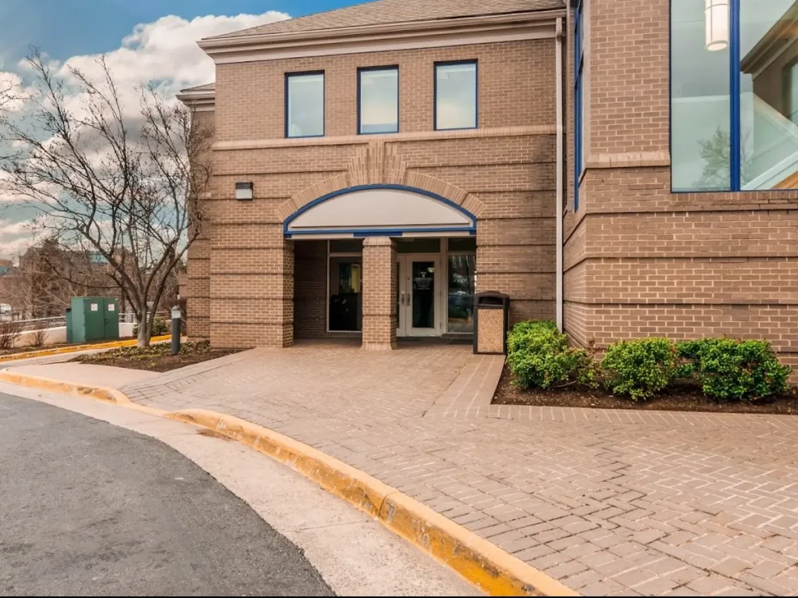 Exterior view of a modern brick dental office entrance with windows, bushes, and paved walkway under a blue sky.