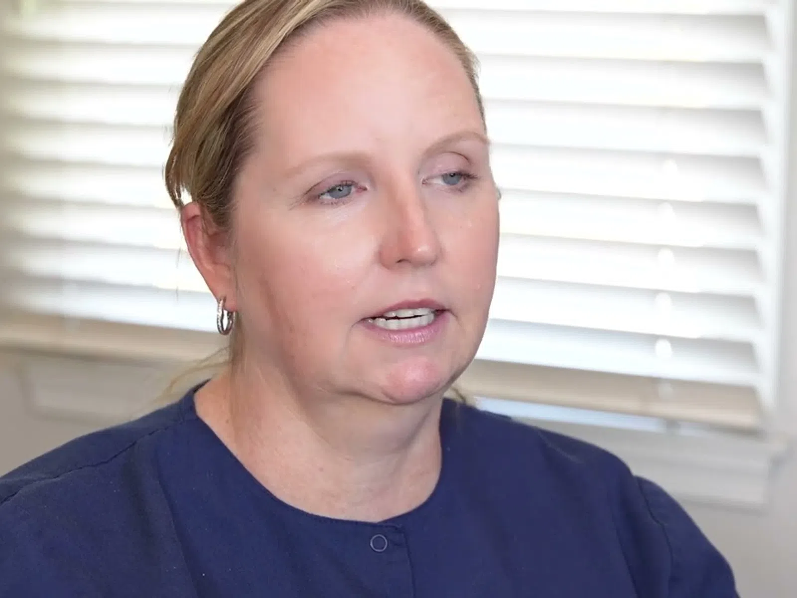 Middle-aged woman in navy blouse speaking indoors with window blinds in the background