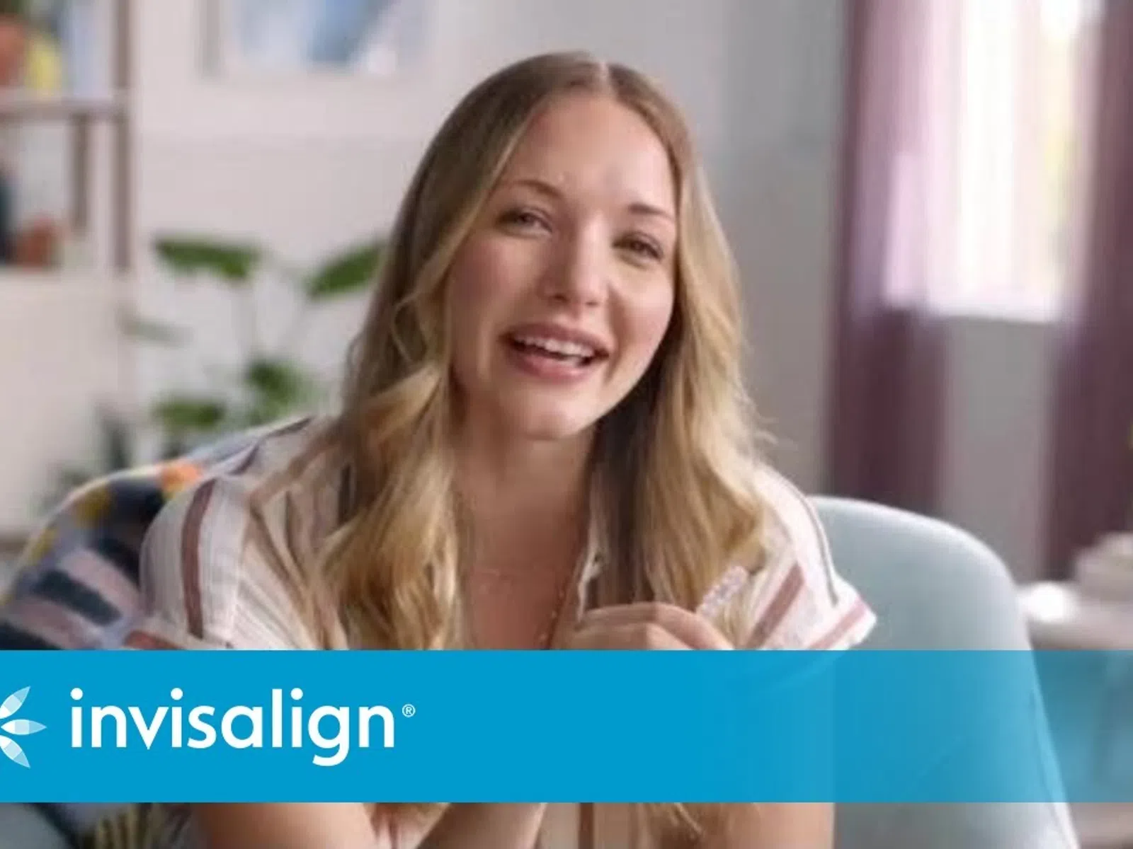 Smiling woman with long blonde hair sitting in a cozy room with a blue Invisalign logo overlay