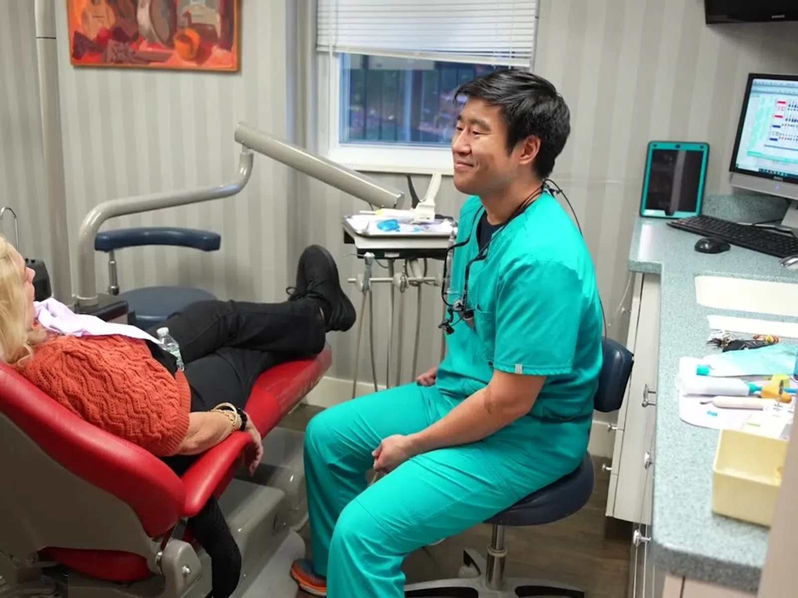 Dentist in teal scrubs talking to female patient reclining in red dental chair in clinic room
