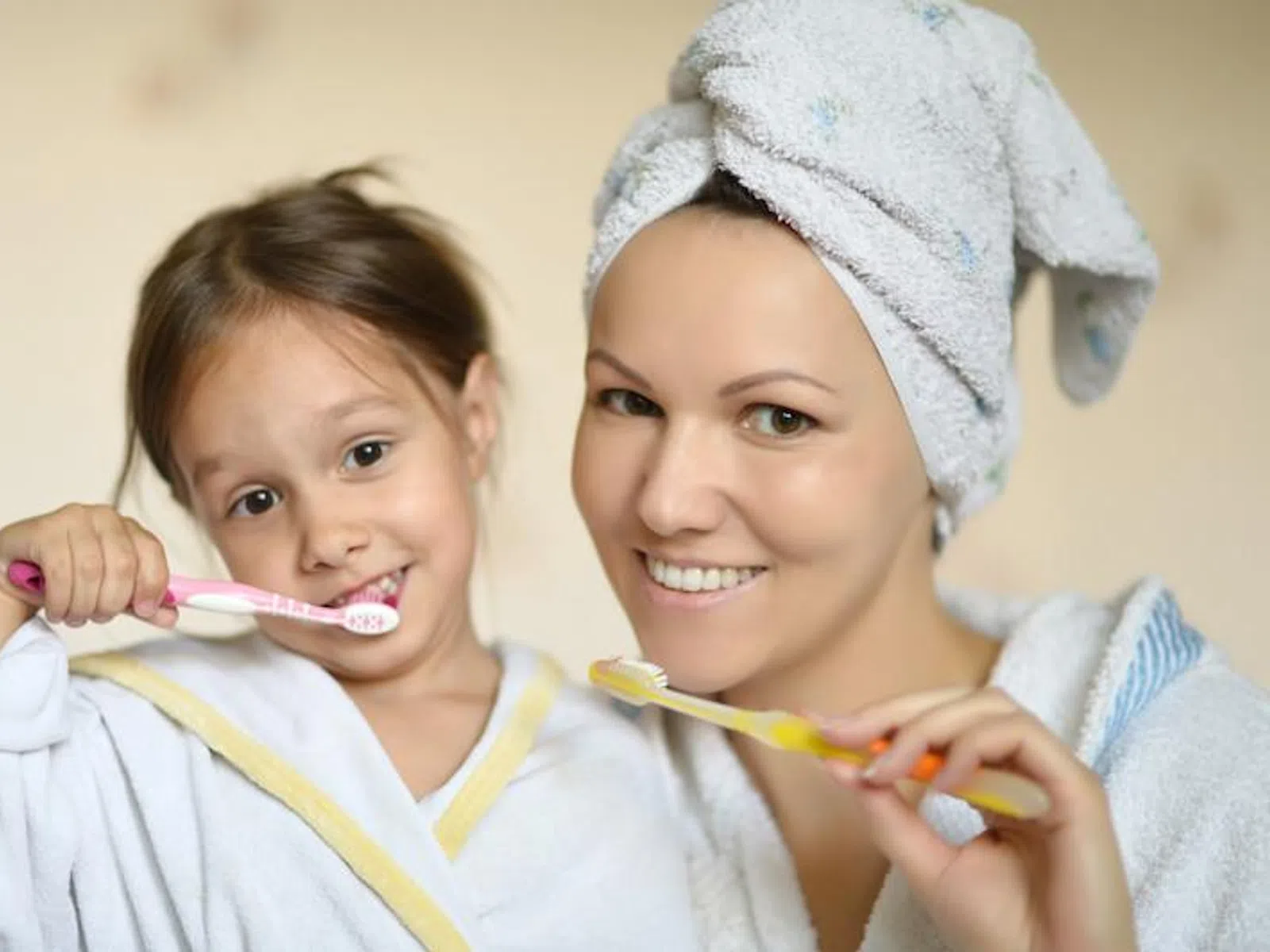 Smiling mother and daughter in bathrobes brushing teeth together at home bathroom.