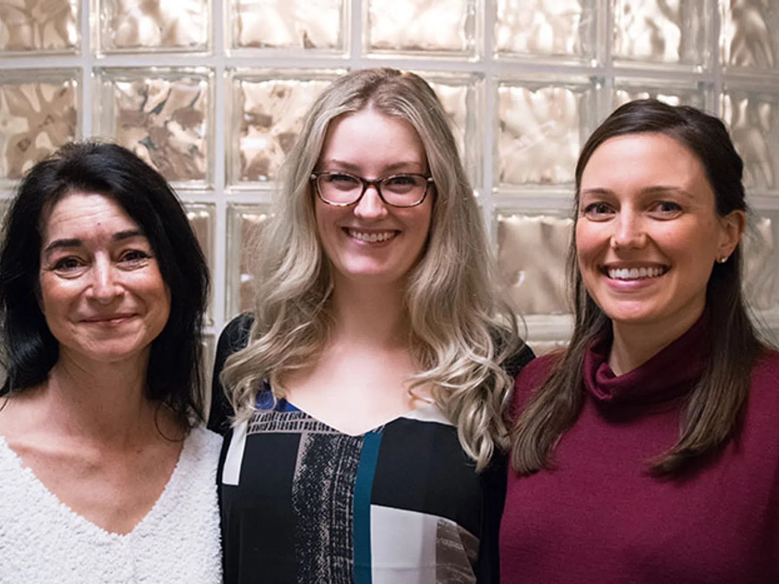 Three smiling women stand side by side against a frosted glass block wall, showcasing diverse hairstyles and casual attire.