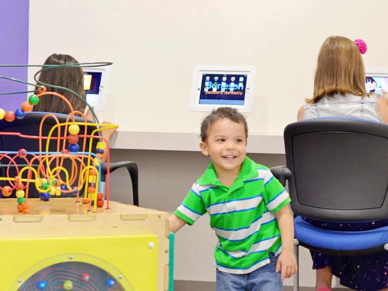 Young boy in green striped shirt playing near colorful bead maze toy in a bright pediatric dentistry waiting room.