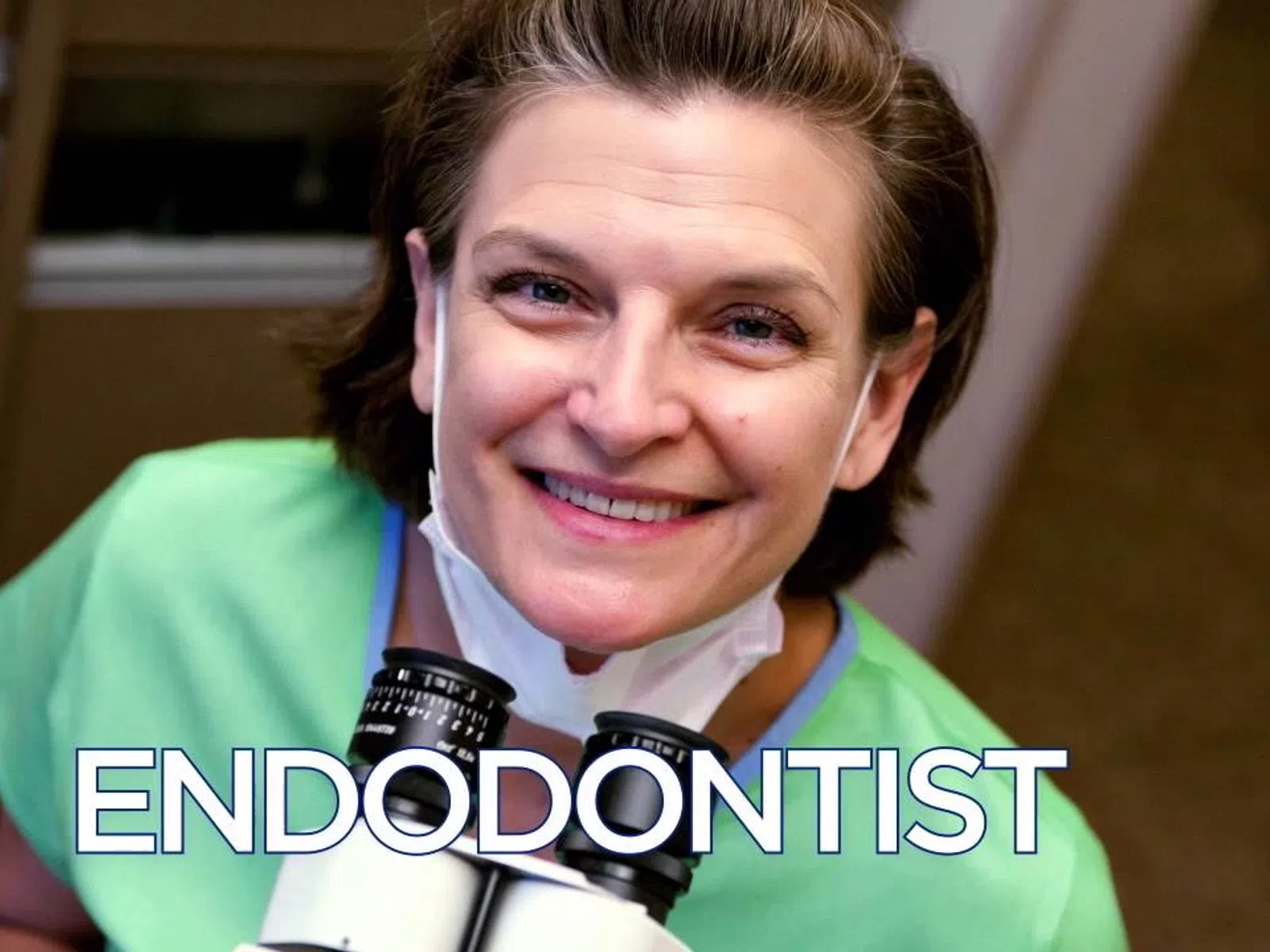 Smiling endodontist in green scrubs with magnifying loupes around neck in dental clinic setting.