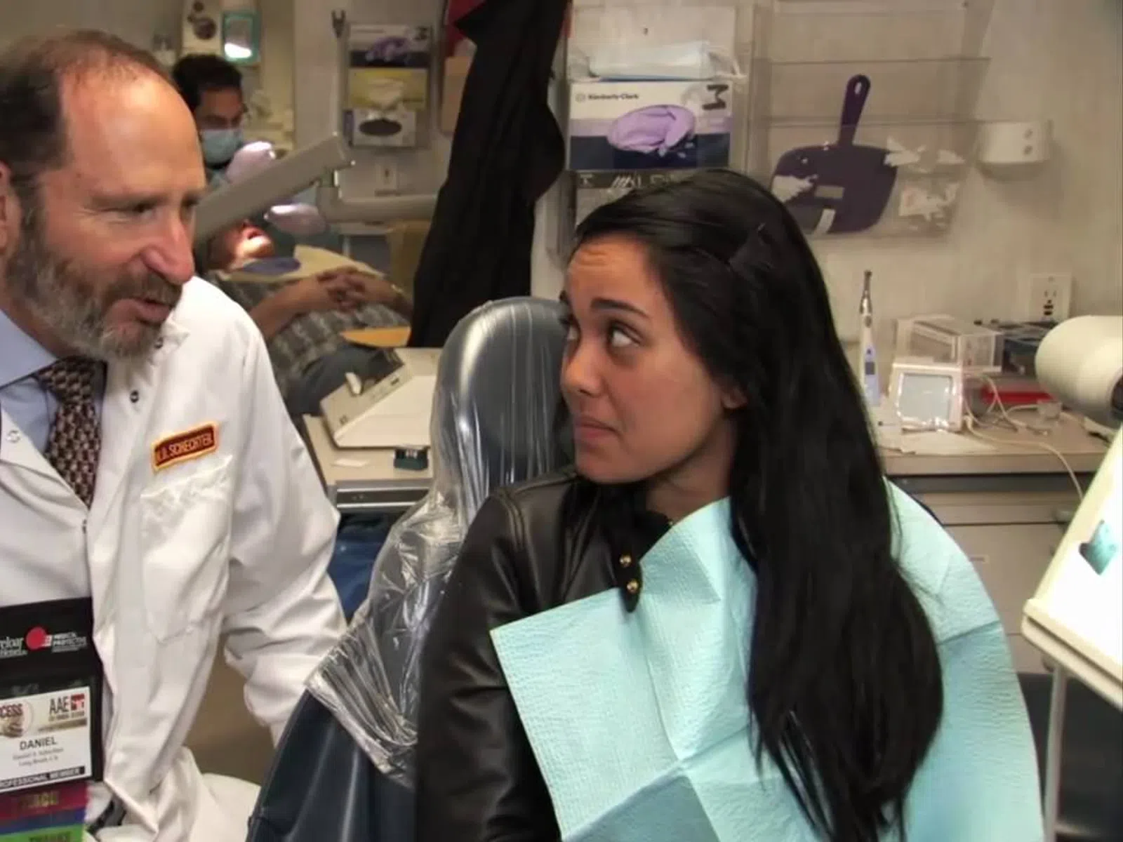 Dentist consulting female patient seated in dental chair inside clinic room with medical equipment.