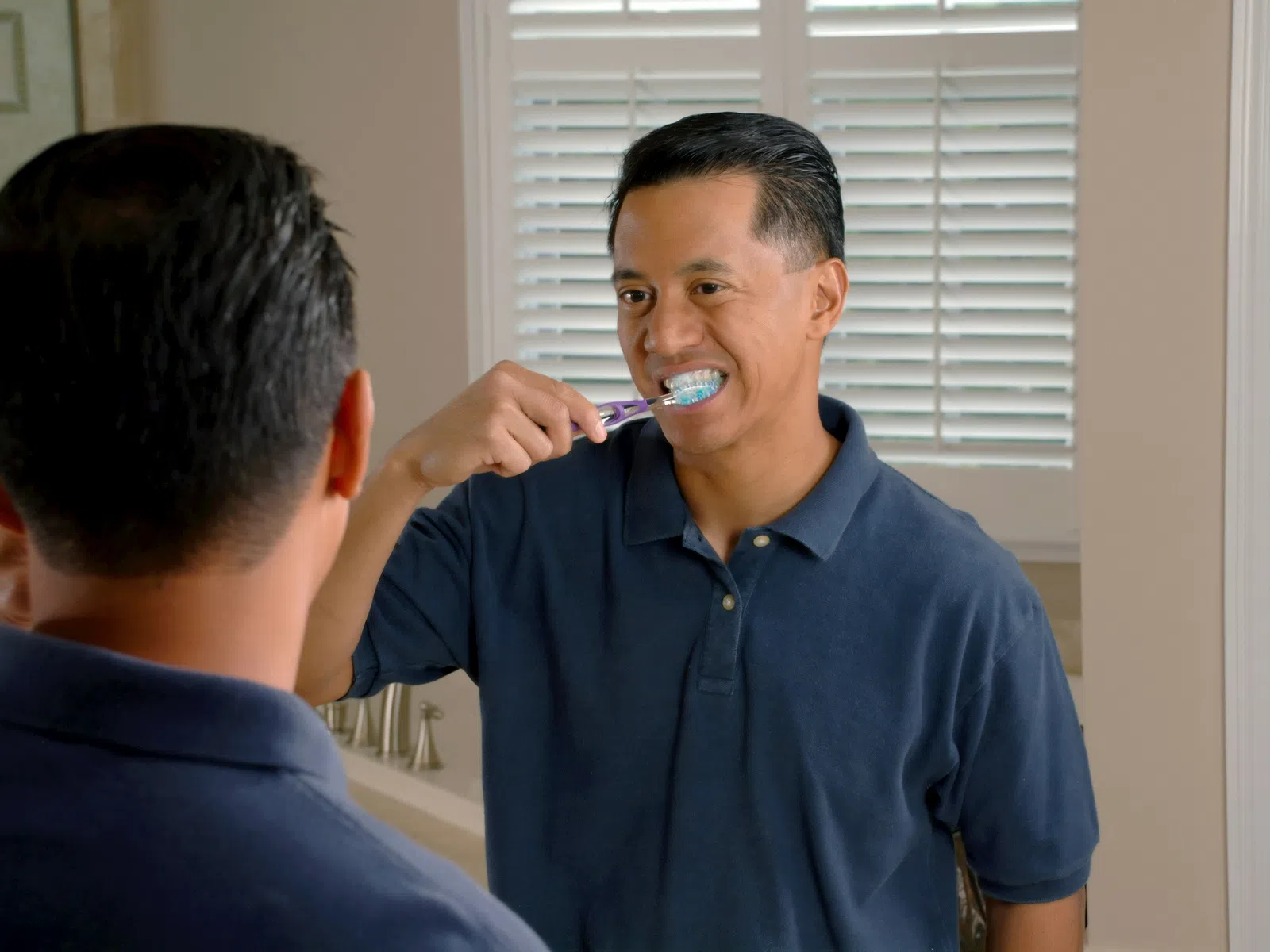 Man wearing navy blue shirt brushing teeth in front of bathroom mirror with white blinds behind.
