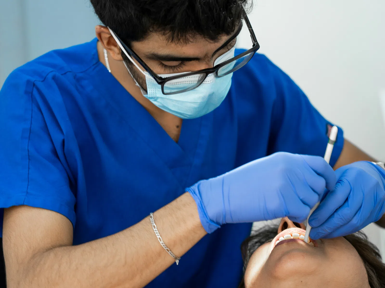 Dentist in blue scrubs and mask examines patient's teeth using dental tools in clinic setting.