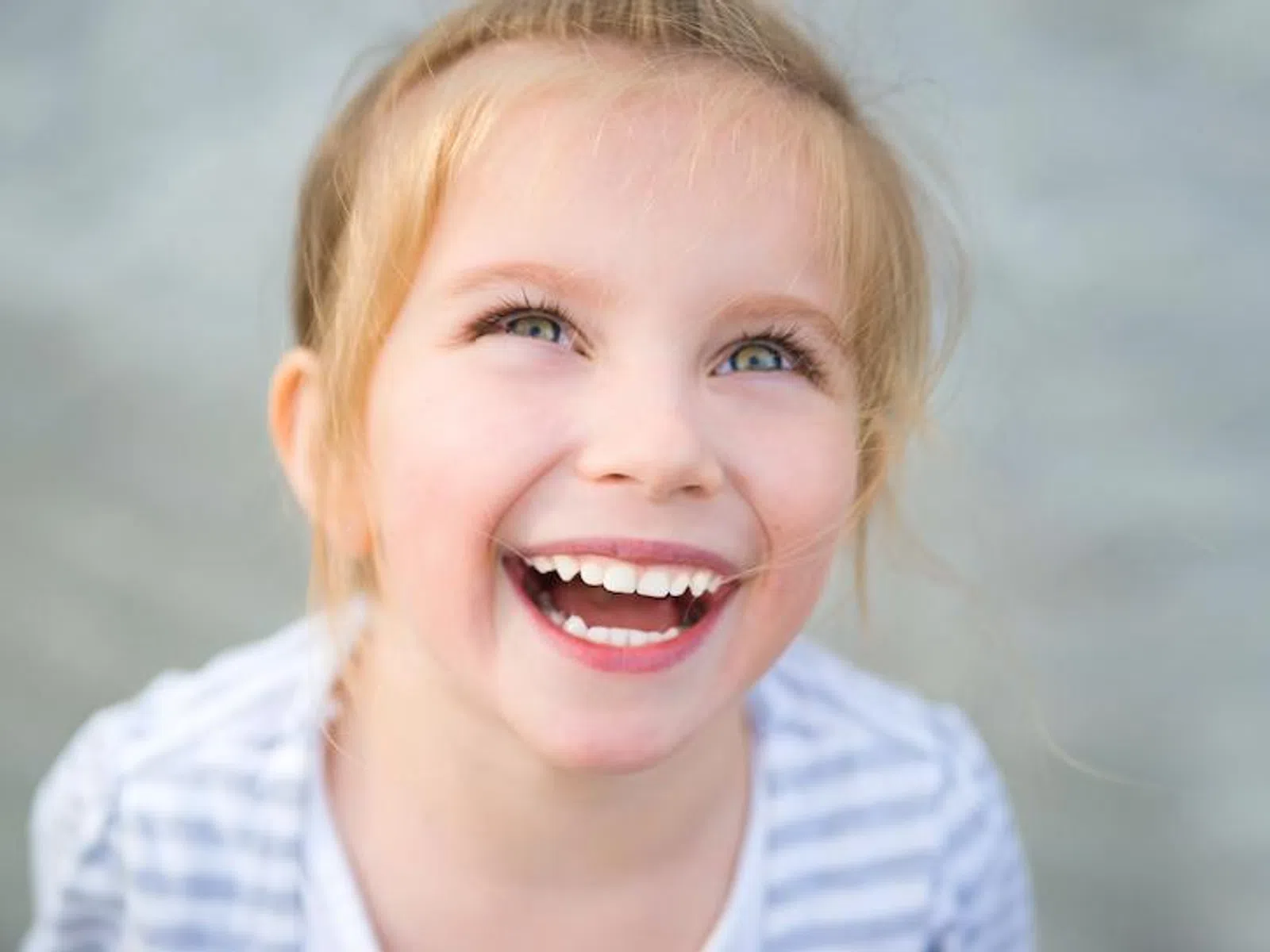 Joyful young girl with blonde hair laughing and looking upwards, wearing a striped shirt against a gray background