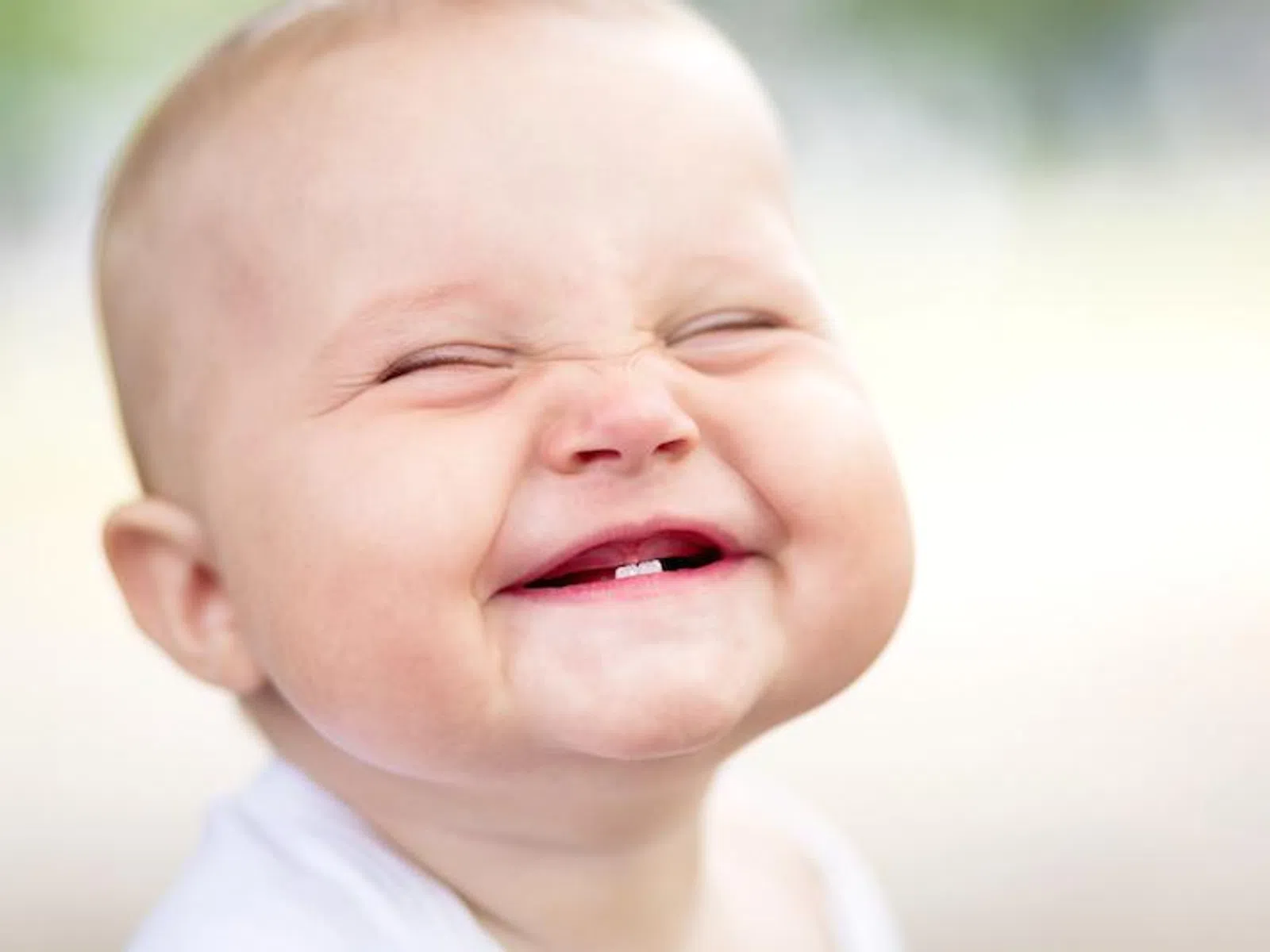 Close-up of a happy baby smiling with eyes closed showing two baby teeth, blurred background.