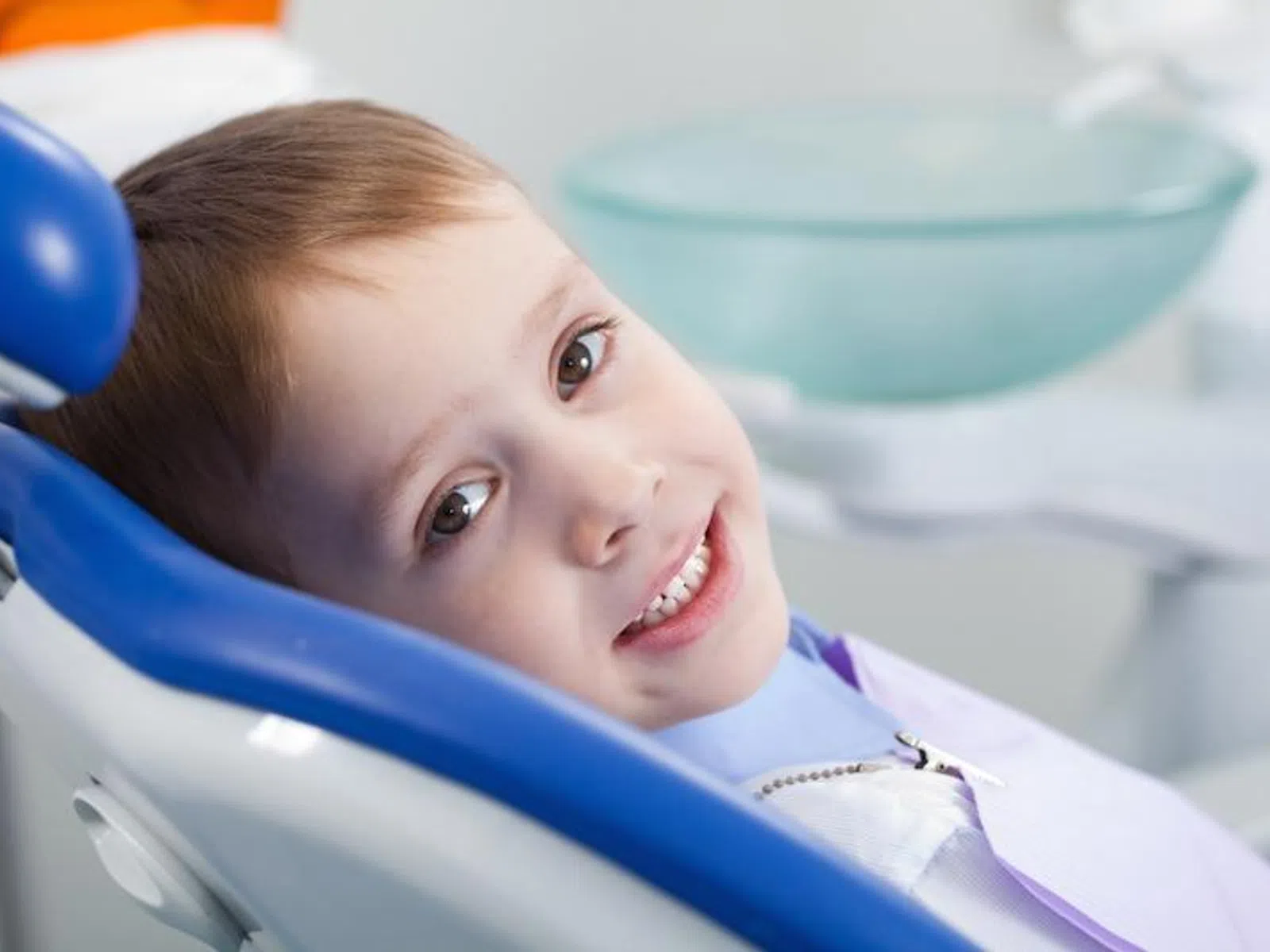 Young boy smiling while sitting in a blue dental chair in a bright dental office.