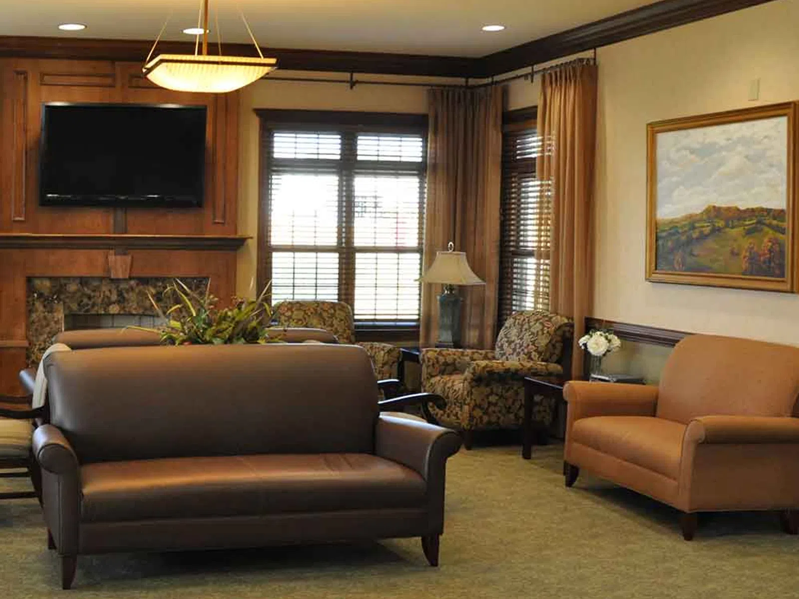 Cozy living room with brown leather sofa, armchairs, fireplace, wall-mounted TV, and large windows with blinds.