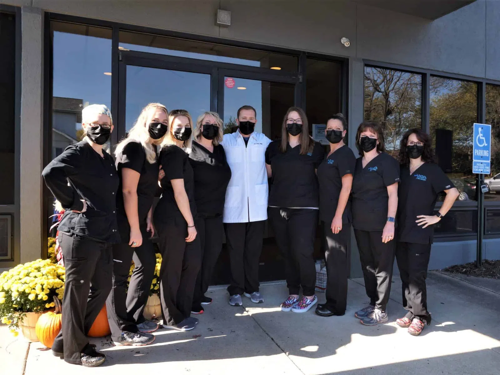 Group of healthcare workers wearing black scrubs and masks standing outside a clinic entrance with autumn decorations.