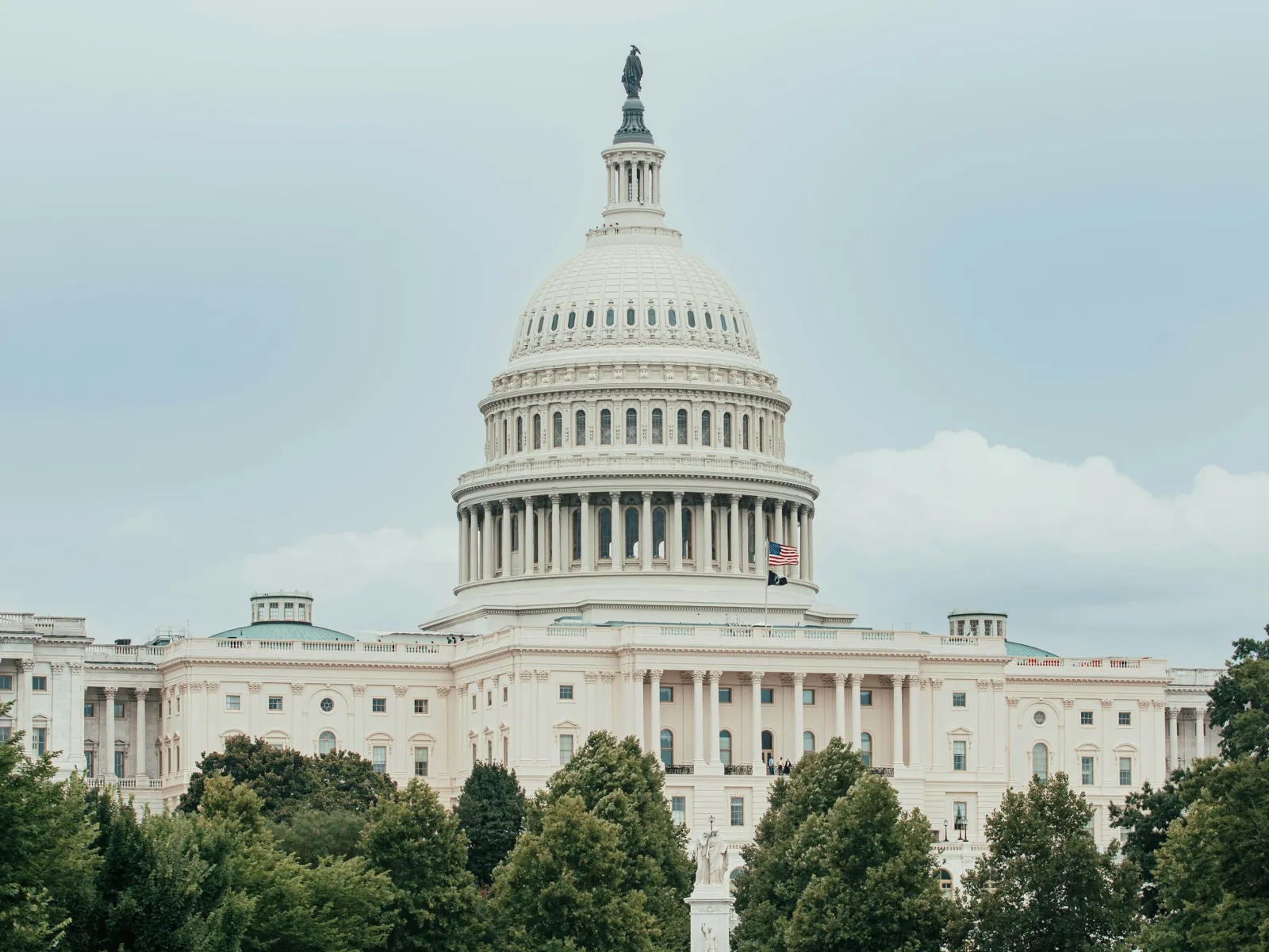 The United States Capitol building with its large dome, American flag, and surrounding trees under a cloudy sky.