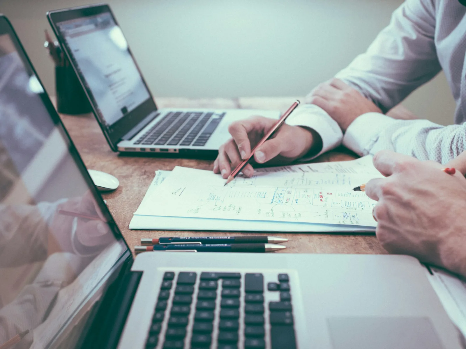 Two professionals collaborating over laptops and documents with handwritten notes at a wooden desk.