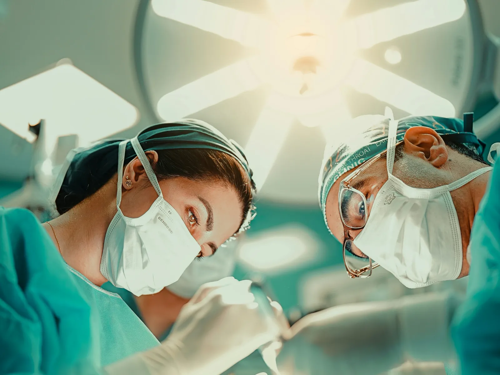Two surgeons wearing masks and scrubs performing surgery under bright operating room lights.