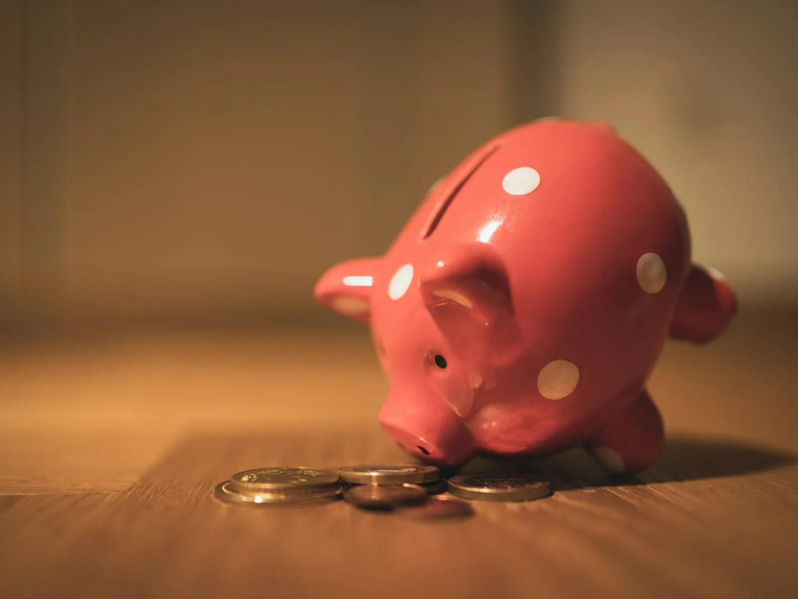Close-up of a pink piggy bank with white polka dots tipped over beside scattered coins on wooden surface.