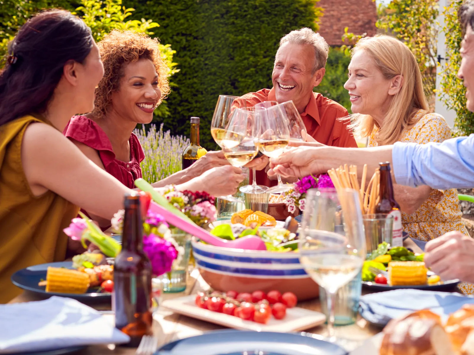 Group of diverse friends enjoying a garden lunch party, toasting with wine glasses around a colorful food-filled table.