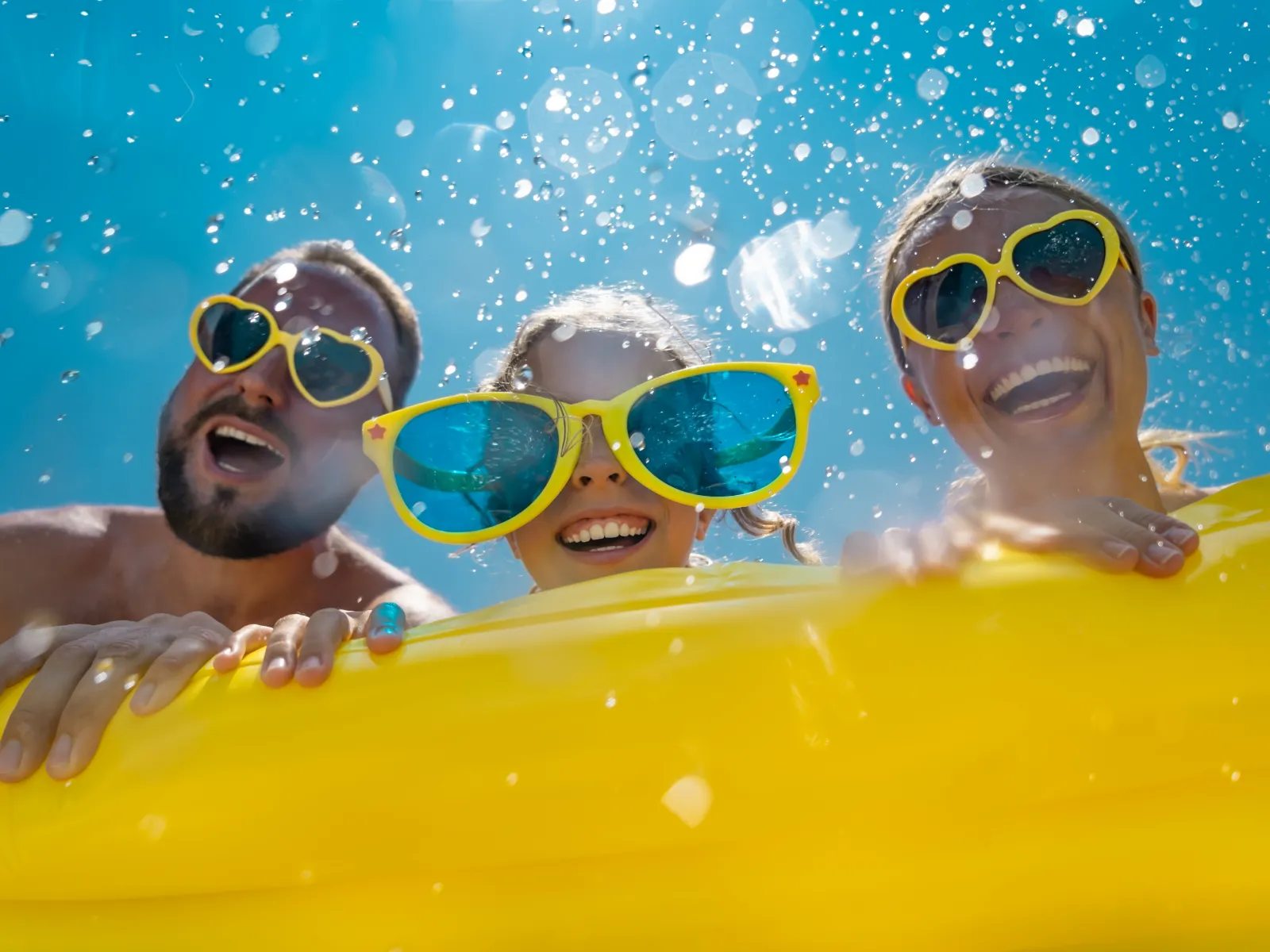 Three people wearing fun sunglasses smiling over a yellow pool float with water splashing and blue sky.